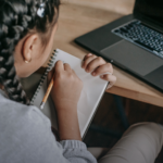 Student writing in a notepad beside an open laptop