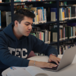 A focused student working on a laptop with an open textbook in a library, studying for the AWS Certified AI Practitioner (AIF-C01) exam