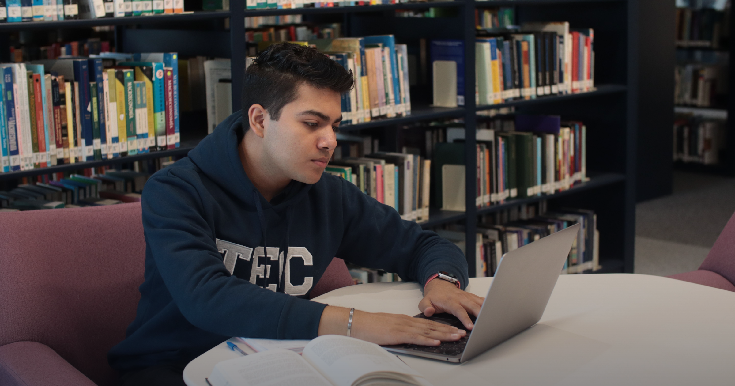 A focused student working on a laptop with an open textbook in a library, studying for the AWS Certified AI Practitioner (AIF-C01) exam