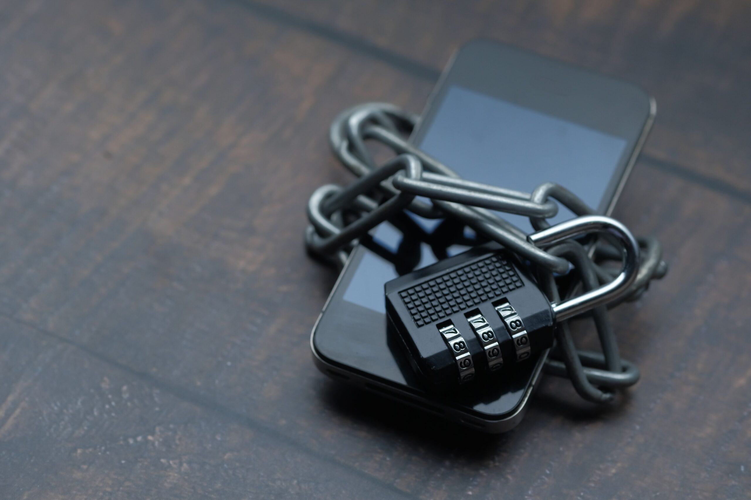 A smartphone wrapped in a metal chain and secured with a combination padlock on a wooden surface